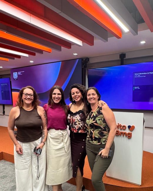 Four women are posing for a group photo in a modern indoor event or office space with large screens and a podium in the background.