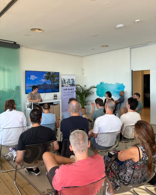 A group of people are seated in chairs indoors, listening to a presentation delivered by a man standing next to a television screen and a promotional banner.