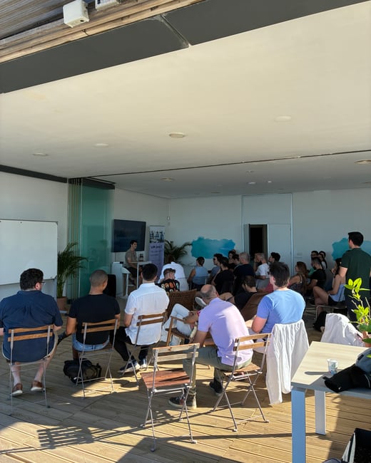 A group of people are seated in an open-sided room, watching a speaker deliver a presentation near a screen and whiteboard.