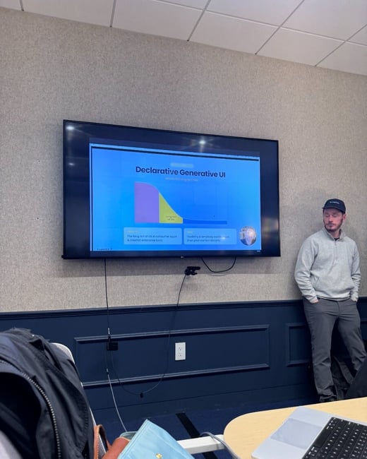 A photograph of a man standing next to a large wall-mounted monitor displaying a presentation slide about 'Declarative Generative UI'. In the foreground, a laptop and personal bags are visible on a table.