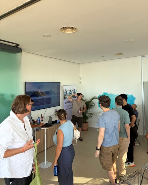 A group of people gathered indoors watching a presentation by a man standing near a TV screen and a banner.