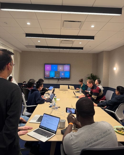 A group of people are gathered around a long conference table in a meeting room, working on laptops and looking towards a large display screen on the wall.