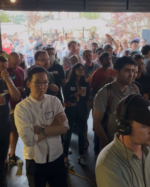 A candid photograph capturing a large, diverse crowd of people gathered indoors at an event, looking toward an unseen focal point, possibly during a presentation or social mixer.