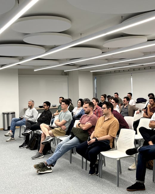A group of people are seated in rows of white chairs, attentively watching a presentation or speaker in a modern indoor space with circular acoustic ceiling panels.