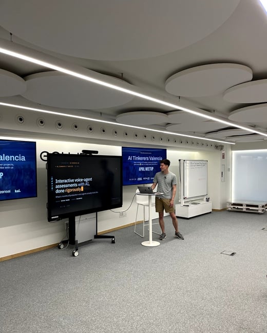 A man stands in a modern event space giving a presentation titled 'Interactive voice-agent assessments with AI, done rigorously' at an AI Tinkerers Valencia meetup.