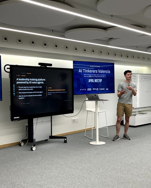 A man is giving a presentation at an 'AI Tinkerers Valencia' meetup, standing next to large screens displaying slides about an AI-powered leadership training platform.