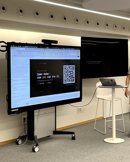 A large digital screen on a rolling stand displays a presentation slide with a QR code, set in an office or conference room next to a high table with a laptop.