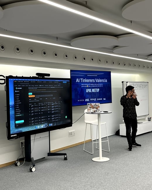 A man is giving a presentation at an 'AI Tinkerers Valencia' meetup, standing next to a large digital display showing a data dashboard.