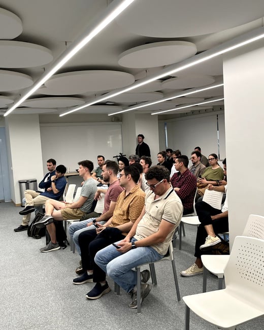 A group of people are seated in a modern meeting room with circular acoustic ceiling panels, appearing to listen to a presentation.