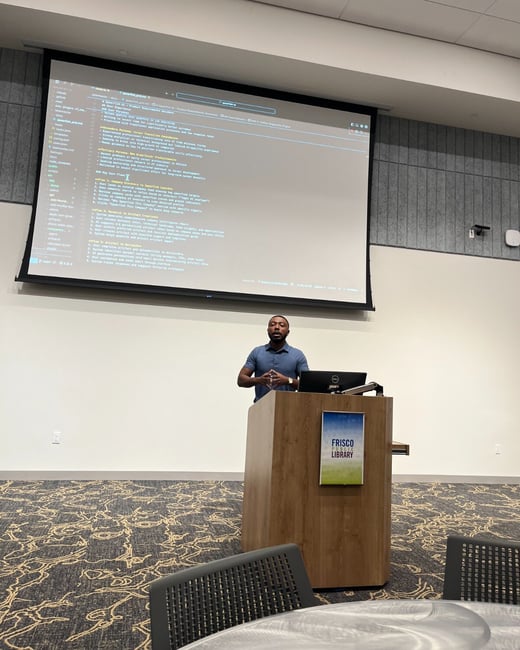 A man is giving a presentation from behind a wooden podium labeled 'Frisco Library' in a meeting room, while a large projection screen displays extensive technical documentation or code.