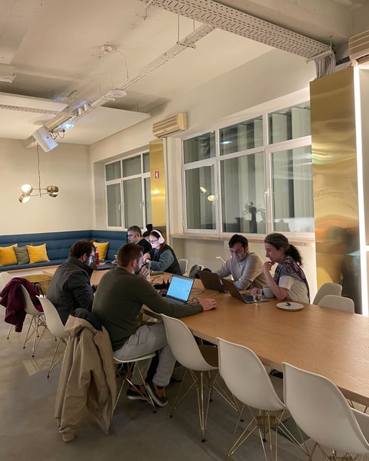 A candid photograph captures five people working on laptops around a long wooden table in a modern, well-lit office or co-working space, characterized by large windows and exposed ceiling infrastructure.