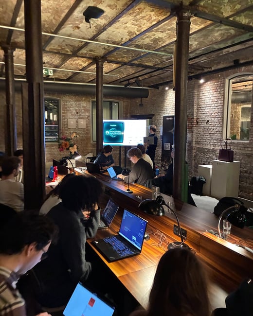 A candid photo taken indoors at a low-light event, likely a hackathon or workshop, showing several people seated along a long wooden counter working intently on their laptops in an industrial-style venue.
