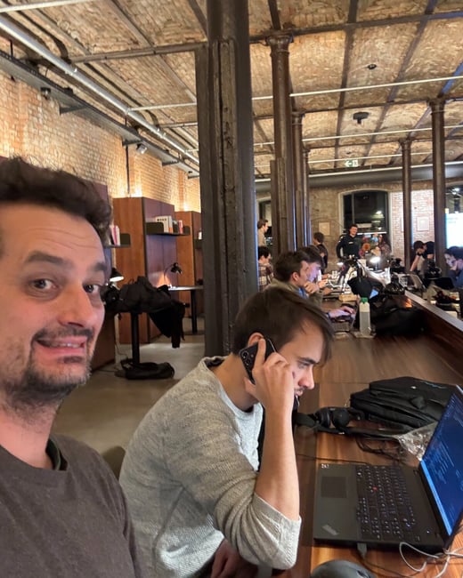 A candid photograph taken in an industrial-style co-working space shows a man smiling directly into the camera while another man next to him is engaged in a phone call and working on a laptop. Several other people are visible working at long desks in the background beneath a high, exposed brick ceiling.