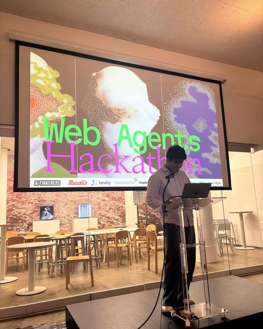 A photograph captures a presenter, a young man, standing at a clear acrylic lectern on a stage, giving a talk during the "Web Agents Hackathon." A large projection screen behind him features neon green and pink text over abstract, dotted imagery, along with sponsor logos.
