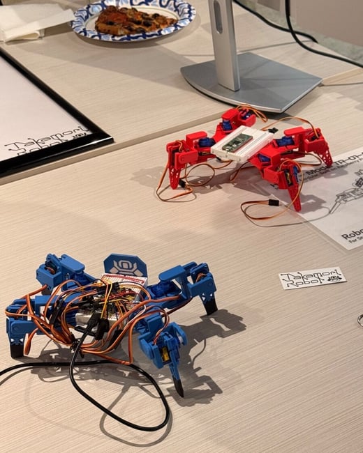 A close-up photograph of a desk showing two small, multi-legged robots (one red, one blue) with exposed wiring and circuit boards, suggesting a robotics project or hackathon setup. The scene also includes a slice of pizza on a paper plate, a beverage can, and the stand of a computer monitor.