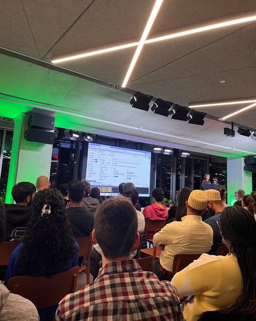 A photograph capturing an audience seated indoors, facing a large projection screen displaying data during a presentation, with modern lighting fixtures and city lights visible through the surrounding windows.