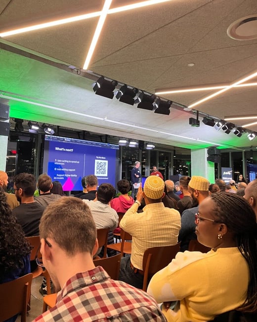 A candid photograph taken from the audience perspective during an indoor presentation event, showing a large crowd seated and facing a screen where two speakers are standing under modern lighting.