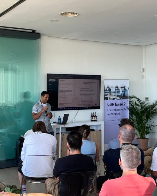 A man stands presenting to an audience in a modern office space next to a screen and a brand banner.