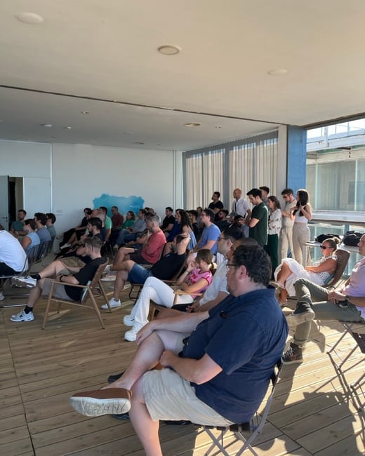 An audience of people sitting and standing in an indoor space, attentively watching a presentation or event.