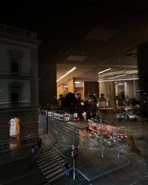 A moody, high-contrast nighttime photograph taken from inside a modern building, capturing a busy city street with red and white trams and pedestrians, overlaid by the reflection of the brightly lit interior and a group of people socializing.