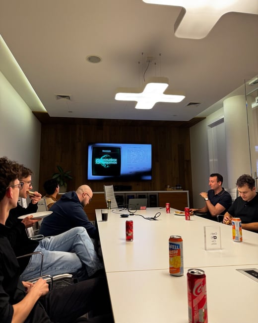 A candid photograph captures six individuals gathered around a white conference table in a modern meeting room, eating snacks and drinking soda while a television screen displays information related to a "WORLDWIDE HACKATHON."