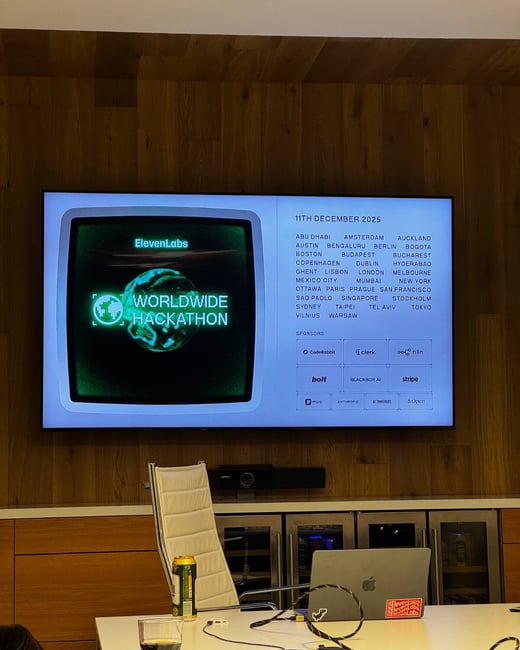 A photograph taken inside a modern office or conference room, showing a large screen mounted on a wood-paneled wall displaying event details for the 'ElevenLabs Worldwide Hackathon' scheduled for December 11, 2025.