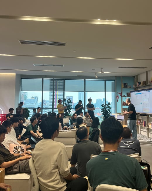 A group of people are gathered in a modern lounge or office space, attentively watching a presentation being delivered by a speaker standing next to a large display screen.