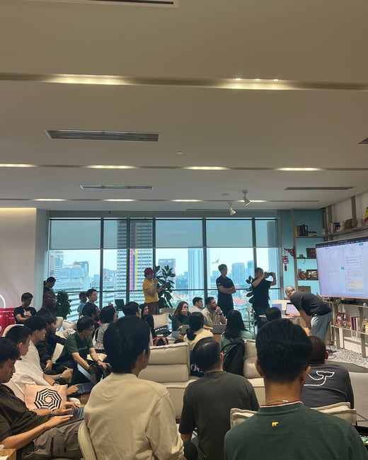 A group of people are gathered in a modern indoor space, likely an office or event venue, watching a presentation or demonstration on a large screen with a city skyline visible through the windows.
