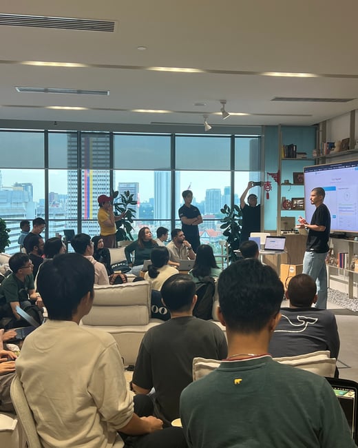 A man stands by a large screen giving a presentation to an audience seated in a modern room with large windows overlooking a city skyline.