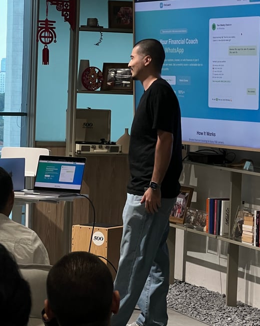 A man in a black t-shirt and light blue jeans is giving a presentation in front of a large screen displaying a financial coaching app called FinCoach.
