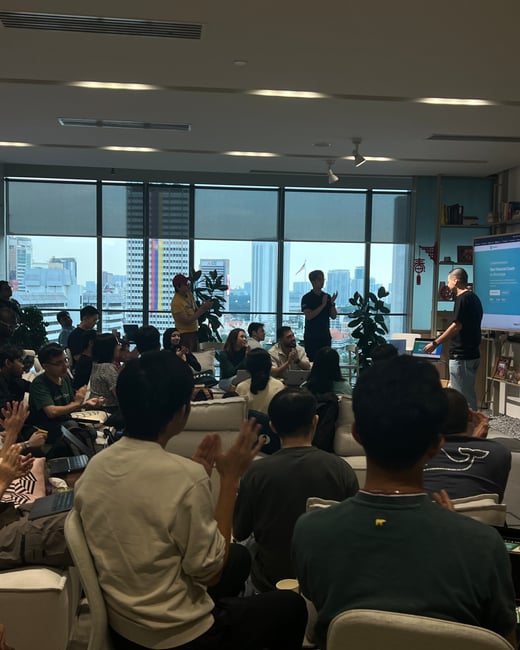 A group of people are gathered in a modern office space with large windows, watching a presentation. The audience is clapping as a man stands next to a large screen displaying a digital service.
