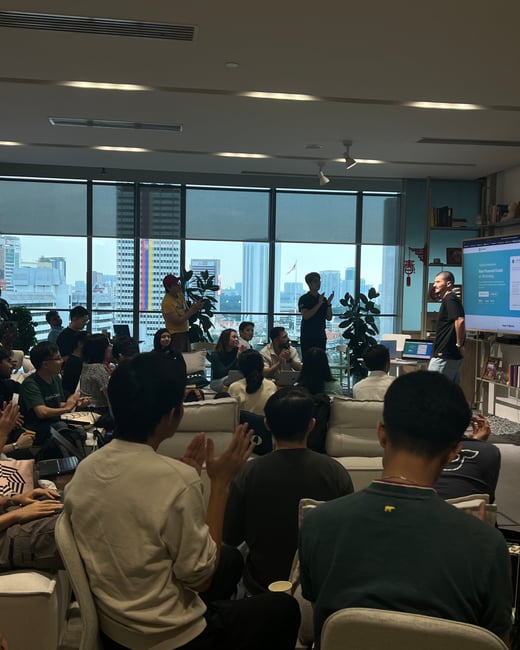 A man is giving a presentation to a seated audience in a modern office space with a large window overlooking a city skyline.