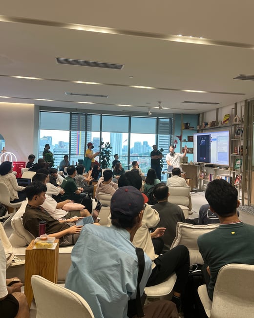 A group of people are gathered in a modern, well-lit indoor space for a presentation. A man stands at the front next to a large screen, gesturing as he speaks to the seated audience.
