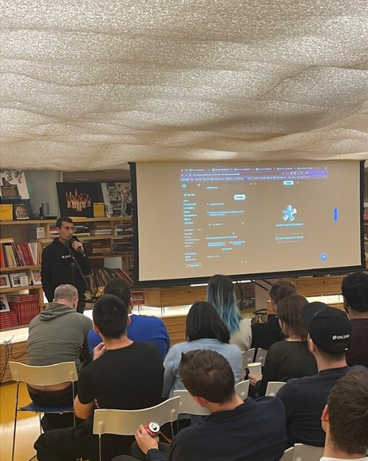 A candid photograph capturing an indoor technology presentation or seminar, showing a speaker holding a microphone next to a large projection screen displaying a software interface, while an audience is seated facing the screen.