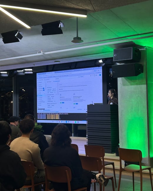 A low-light photograph captures a technical presentation taking place indoors, showing an audience seated, facing a large screen displaying code and application data, with a presenter standing at a podium illuminated by green accent lighting.