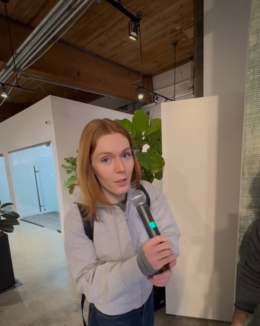 A young woman with reddish hair, wearing a grey zip-up hoodie and dark jeans, stands indoors holding a microphone, appearing to be speaking or giving an interview.