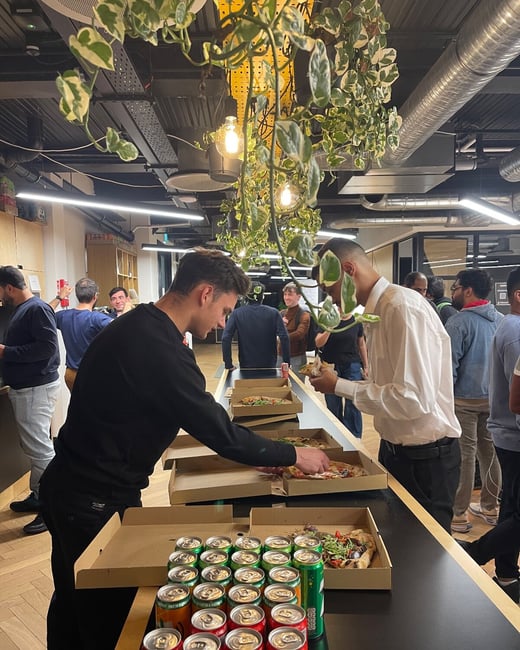 This is a photograph of a casual office gathering where attendees are serving themselves pizza and canned sodas from a long table, set beneath hanging plants and industrial lighting.