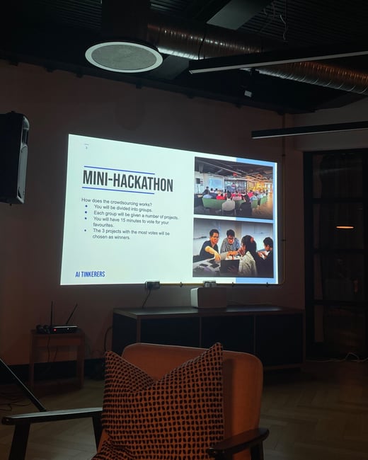 A photograph taken indoors captures a presentation slide projected onto a wall in a dimly lit event space, detailing the rules for a "MINI-HACKATHON." The foreground features a comfortable chair with an orange and black patterned pillow, suggesting the viewer's perspective from the audience.