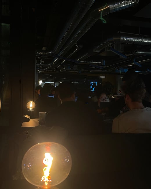 A dark, atmospheric photograph of people seated indoors, featuring an industrial ceiling with exposed ductwork and a prominent, glowing Edison-style light bulb in the immediate foreground.