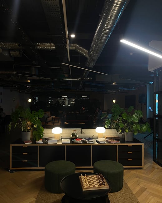 A dimly lit, modern interior photograph of a lounge or common area featuring a low cabinet with potted plants and two glowing table lamps, set against a backdrop of exposed industrial ceiling ducts. In the foreground, a small round table holds a wooden chessboard.