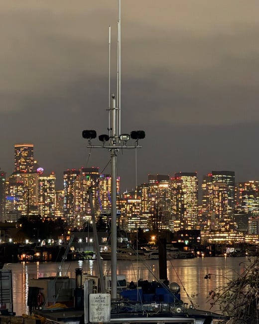A nighttime photograph captures a moored vessel or houseboat in the foreground, featuring a tall mast with lights, set against the backdrop of a brightly illuminated city skyline across the water.