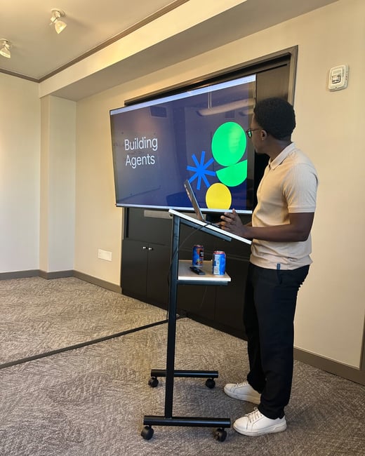 A photograph capturing a man giving a presentation in an indoor meeting room, standing next to a mobile stand while facing a large screen that displays the title slide, "Building Agents."