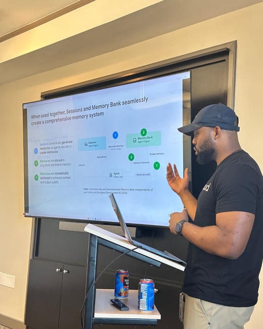 A man wearing a black t-shirt and baseball cap is giving a technical presentation in an office setting, standing next to a large screen displaying a flowchart about a comprehensive memory system.