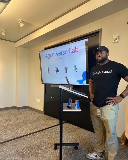 A man wearing a black Google Cloud t-shirt and a baseball cap is presenting in a meeting room, standing next to a large screen displaying the title "Agentverse Lab."