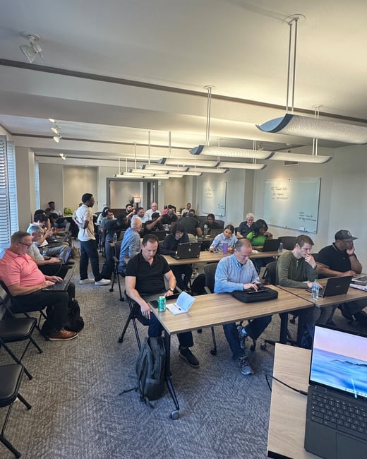 A photograph capturing a large group of people attending an indoor training session or workshop, seated at tables with laptops open in a modern classroom setting.