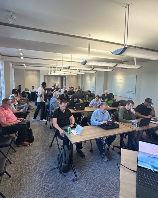 A candid photograph taken indoors showing a large group of people attending a workshop or training session, seated at long tables with laptops open in a brightly lit room.