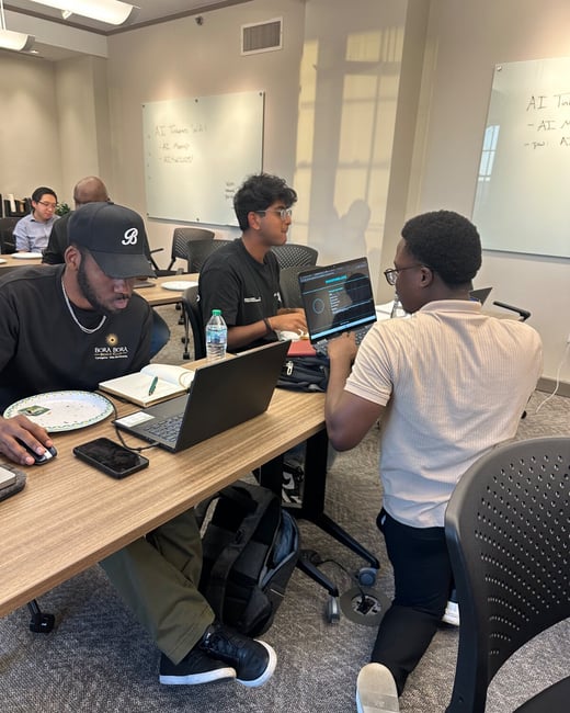 A candid indoor photograph capturing a group of men working collaboratively around a table during a technical workshop or class, with several laptops open and whiteboards visible in the background containing handwritten notes about 'AI Themes'.