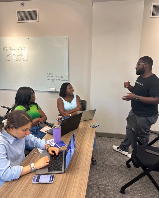 A candid photograph captures four people—three women seated at a wooden table with open laptops and one standing man—engaged in a discussion in a modern meeting room setting.