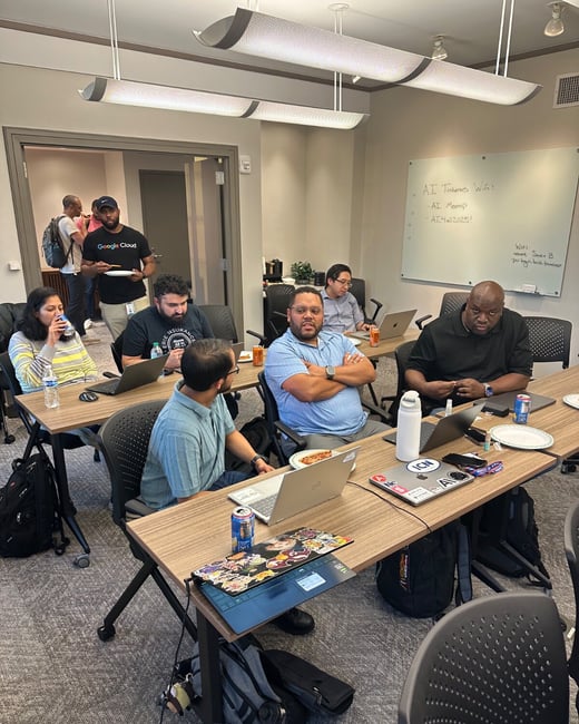 A photograph captures a casual tech meeting or workshop taking place indoors, showing a diverse group of people seated at folding tables with laptops open, drinks, and pizza, while a whiteboard displays Wi-Fi login details for an 'AI Tinkers' event.