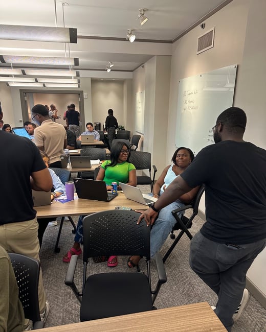 A candid photograph taken inside a modern classroom or workspace where a diverse group of people is gathered around tables with laptops open, suggesting a workshop or collaborative meeting.